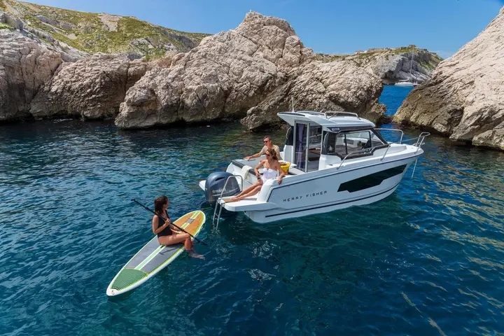 Jeanneau Merry Fisher 605: boat moored in a rocky cove with people in the cockpit and a paddleboarder at the stern