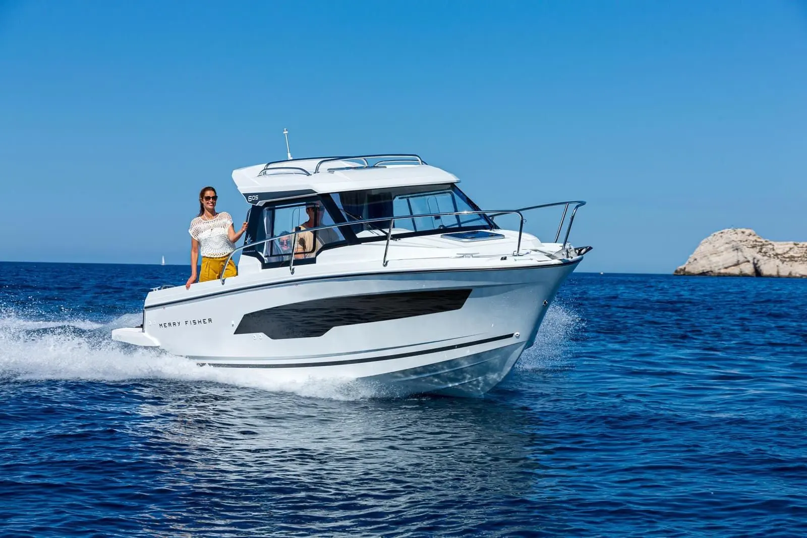 Jeanneau Merry Fisher 605: wheelhouse motorboat cruising on open water with a woman on the side deck, rocky islet behind