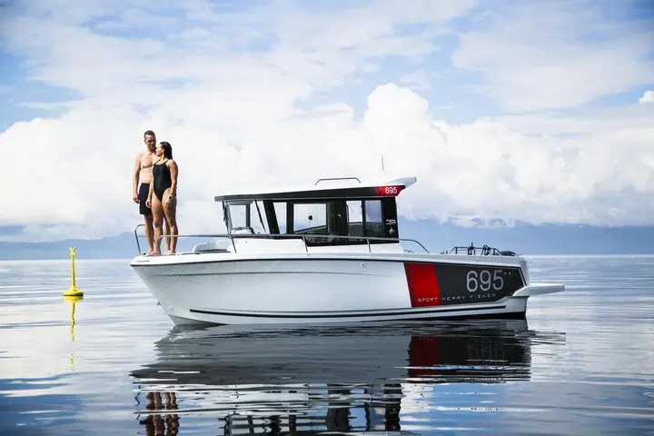Jeanneau Merry Fisher 695 Sport: couple standing on the foredeck of the white boat on calm water, wheelhouse and outboard bracket visible