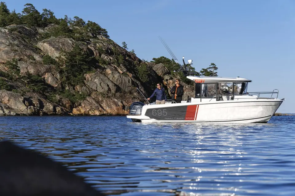 Jeanneau Merry Fisher 695 Sport: side view of white boat near a rocky wooded island, two people fishing aft of the wheelhouse