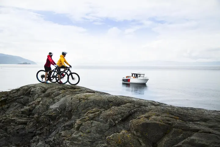 Jeanneau Merry Fisher 695 Sport: two cyclists on a rocky shore overlooking calm water, with the white boat at anchor offshore