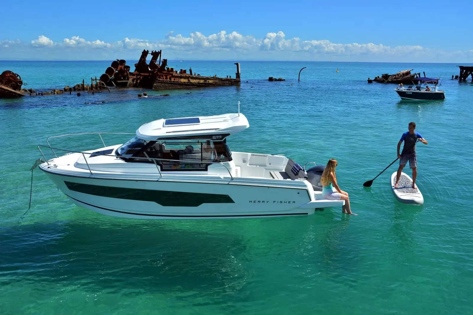 Jeanneau Merry Fisher 695: boat at anchor in clear turquoise water, woman sitting on the bathing platform, paddleboarder alongside