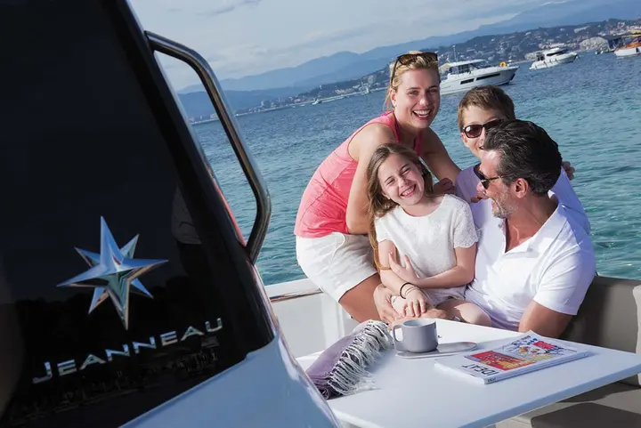 Jeanneau Merry Fisher 695: family of four laughing around a white cockpit table beside the wheelhouse, sea and moored boats behind