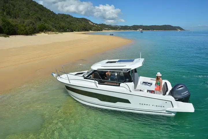 Jeanneau Merry Fisher 695: white cabin boat at anchor in shallow clear water beside a sandy beach and wooded hills