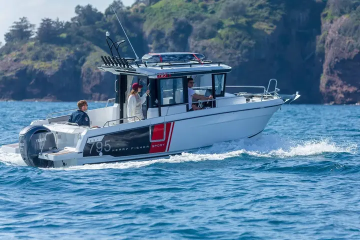 Jeanneau Merry Fisher 795 Sport: side view underway past a green rocky headland, three people aboard in the cockpit and wheelhouse