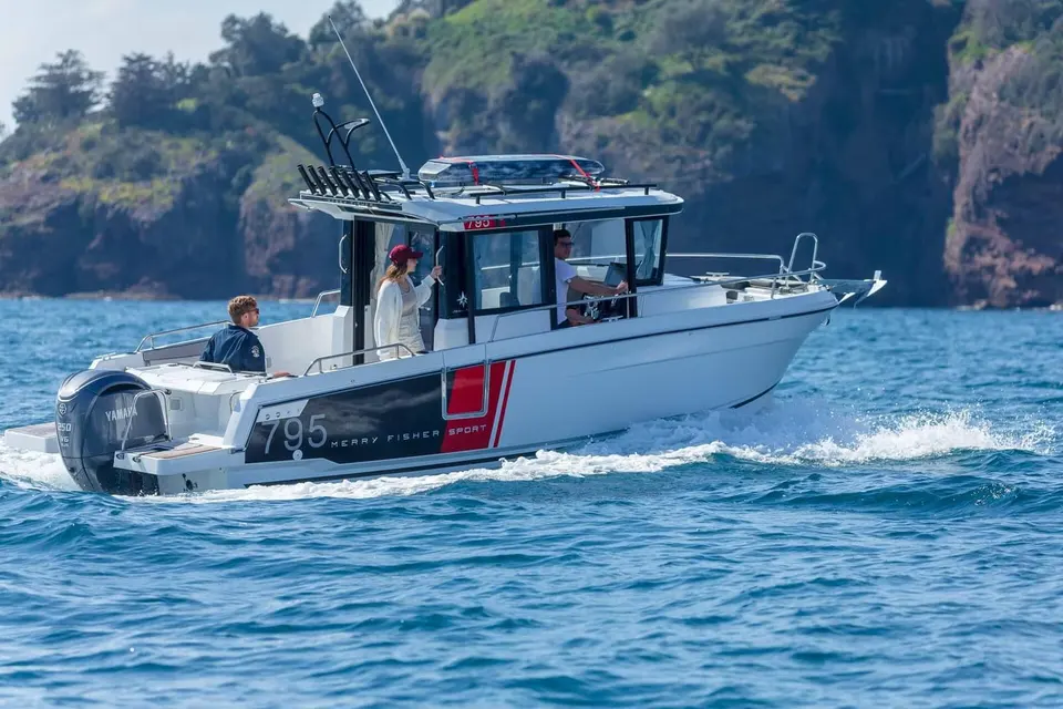 Jeanneau Merry Fisher 795 Sport: side view underway past a green rocky headland, three people aboard in the cockpit and wheelhouse