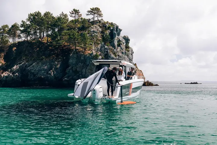 Jeanneau Merry Fisher 895 Sport Série 2: stern view at anchor near a rocky wooded islet, twin outboards, person in wetsuit with foil board