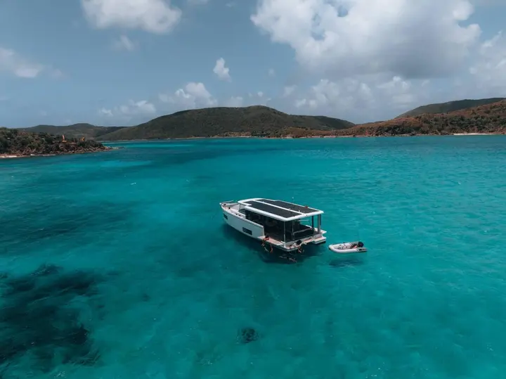 Jeanneau Sea Loft 480: aerial view at anchor in turquoise bay, solar panels on roof, tender alongside, green hills behind