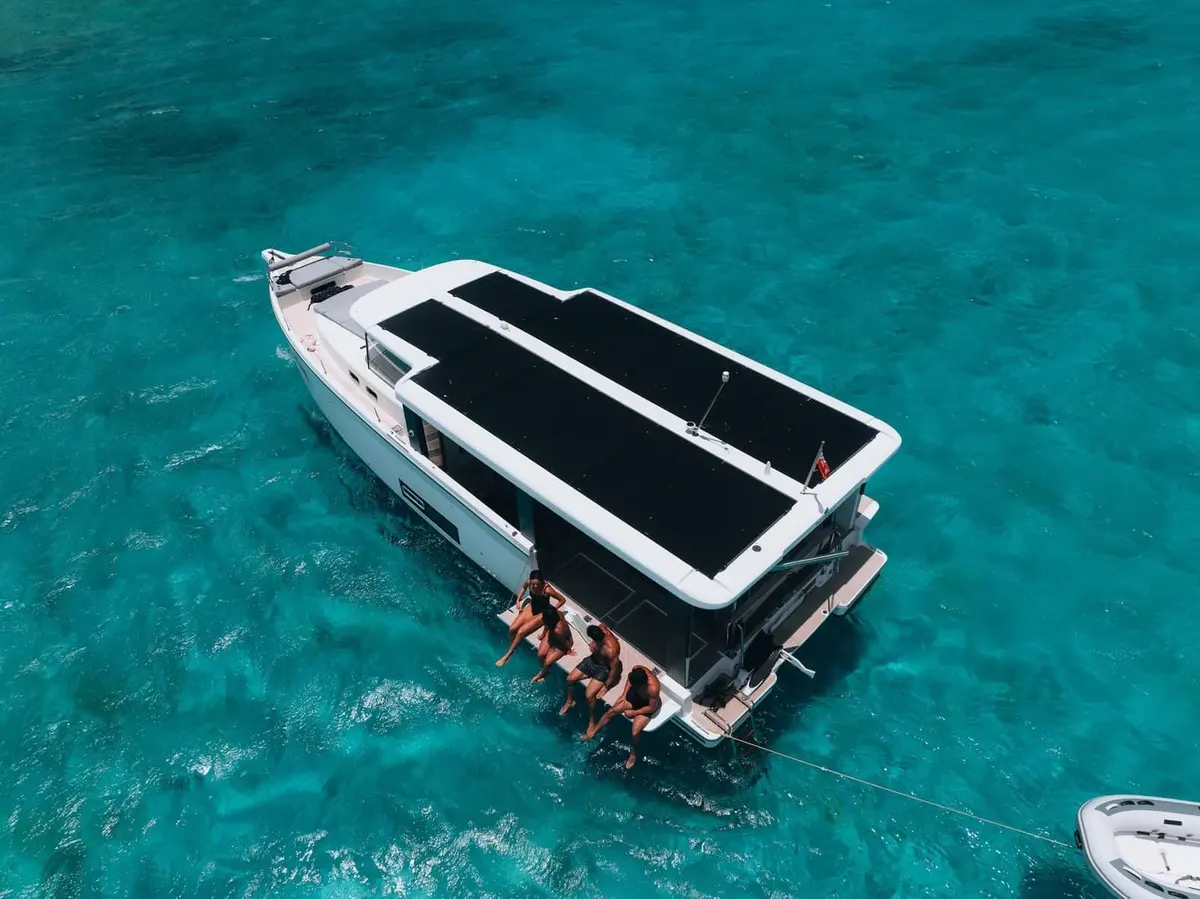 Jeanneau Sea Loft 480: aerial view at anchor in turquoise water, solar panels covering the roof, four people sitting on the bathing platform