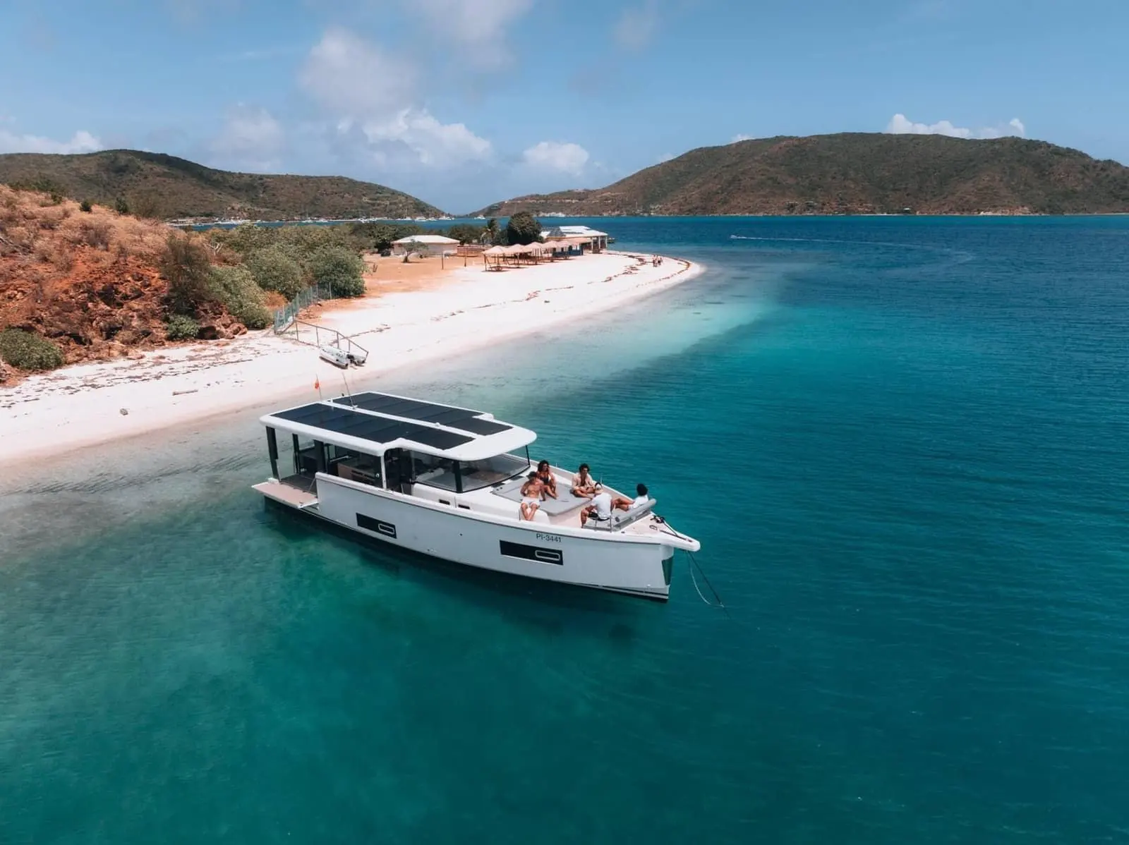 Jeanneau Sea Loft 480: moored off a white sandy beach with hills behind, guests relaxing on the forward seating area