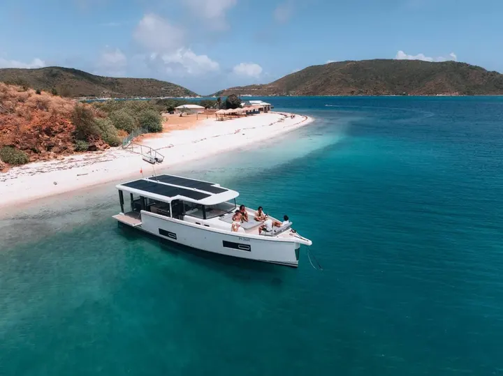 Jeanneau Sea Loft 480: moored off a white sandy beach with hills behind, guests relaxing on the forward seating area