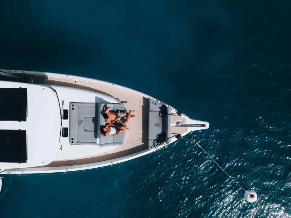 Jeanneau Sea Loft 480: overhead view of the foredeck with grey sunpad, two people lounging, anchor line leading to a buoy