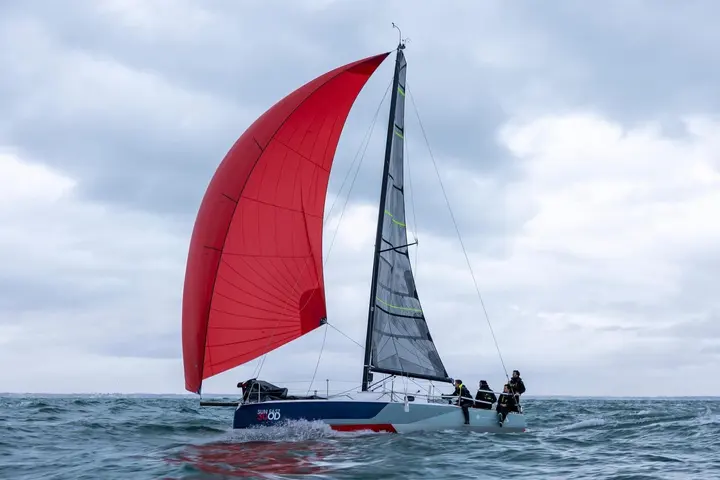 Jeanneau Sun Fast 30 OD: full view under red spinnaker and dark mainsail beneath grey clouds, crew sitting aft