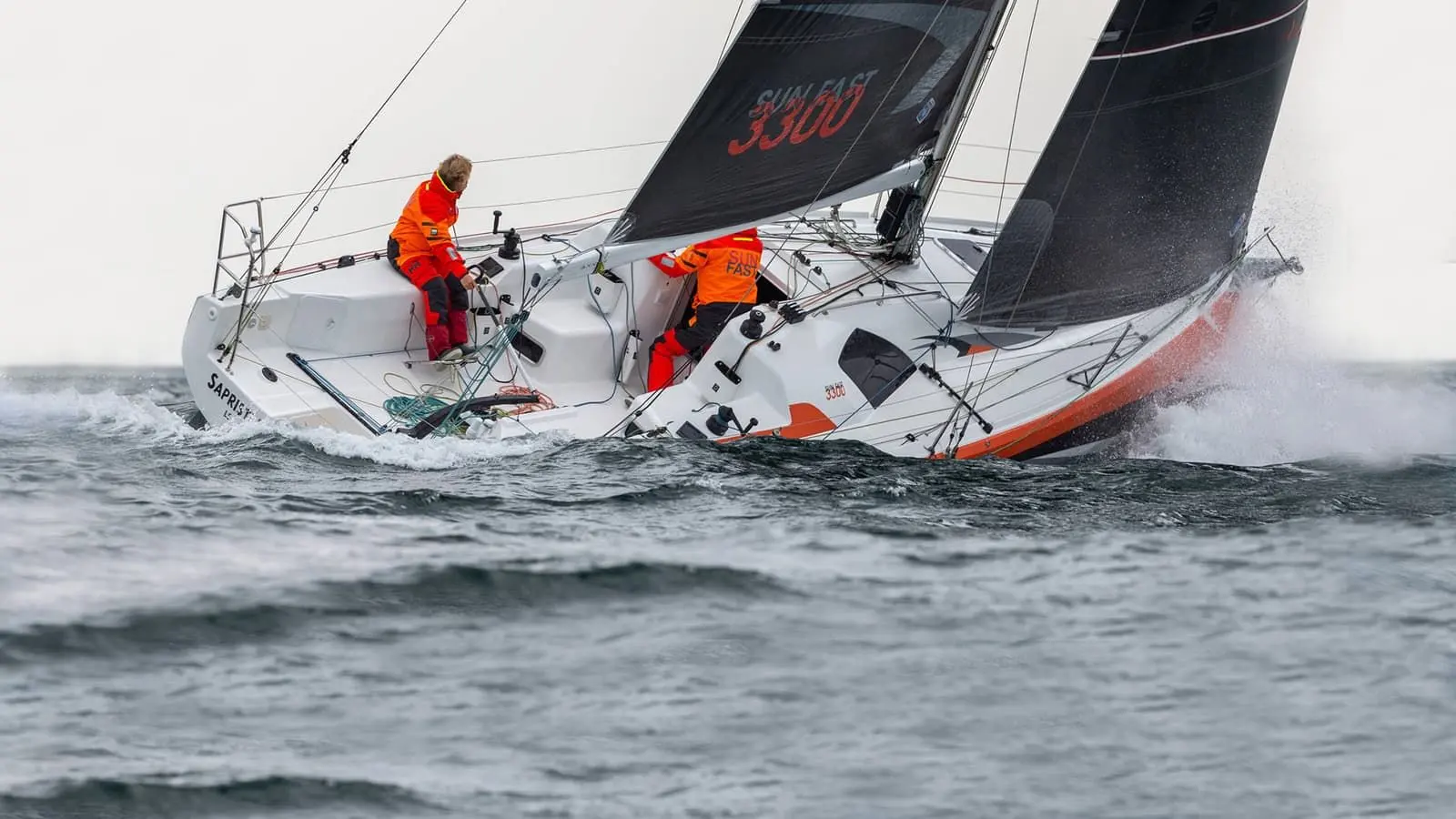 Jeanneau Sun Fast 3300: under sail heeled in choppy grey sea, two crew in orange foul-weather gear on deck