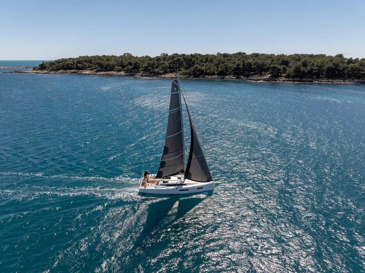 Jeanneau Sun Odyssey 350: aerial view of the yacht under sail with dark grey sails, passing a wooded island