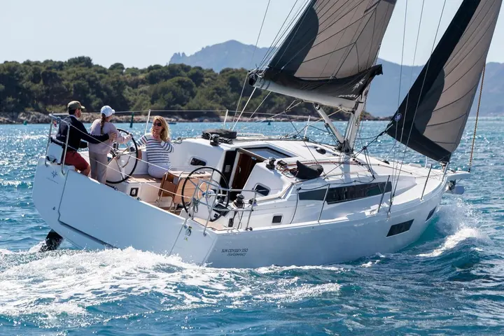 Jeanneau Sun Odyssey 350: under sail from the stern quarter, three people at the twin wheels in the cockpit, wooded coast behind
