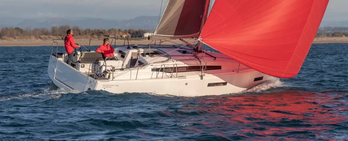 Jeanneau Sun Odyssey 380: white sailing yacht under sail with red gennaker, two crew in red jackets at the twin helms