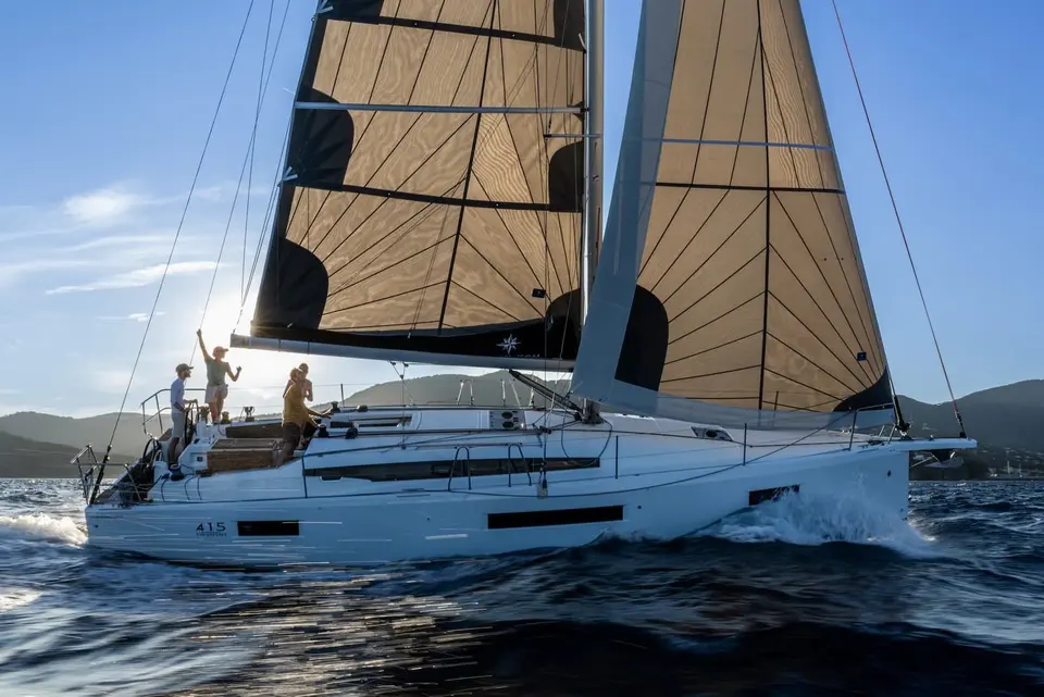 Jeanneau Sun Odyssey 415: under sail with tan and black sails, crew on deck at sunset, hills behind