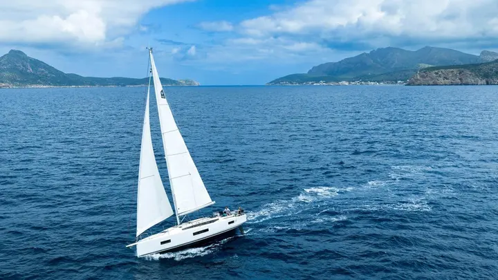Jeanneau Sun Odyssey 455: aerial view under sail with mainsail and headsail set, crew in cockpit, mountains behind