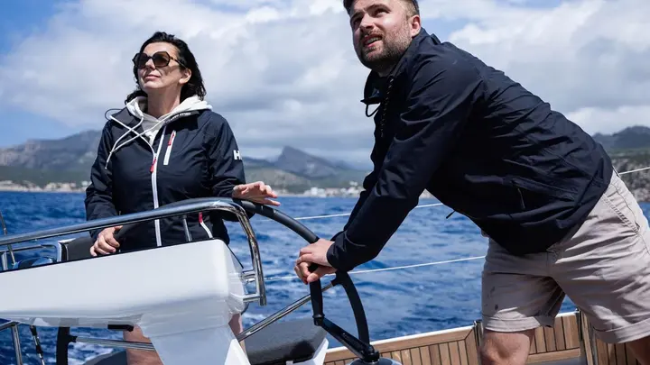 Jeanneau Sun Odyssey 455: two people at the helm under sail, one steering at the wheel, coastline behind