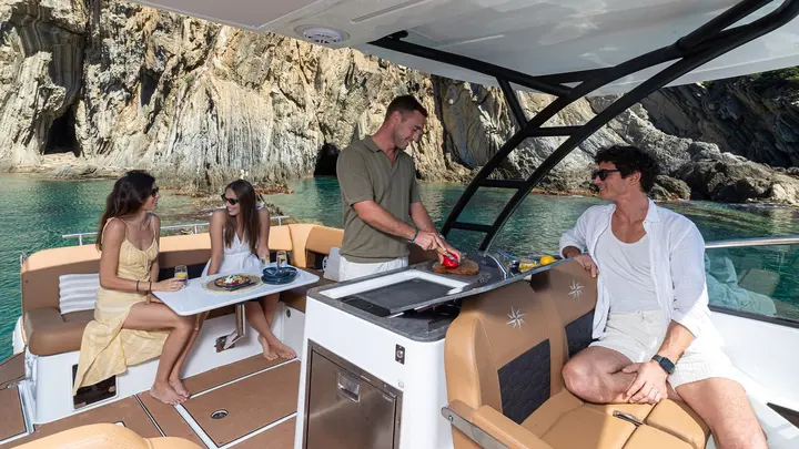 Jeanneau TH33: shaded cockpit with outdoor galley counter, table, tan seating and four people beside rocky cliffs