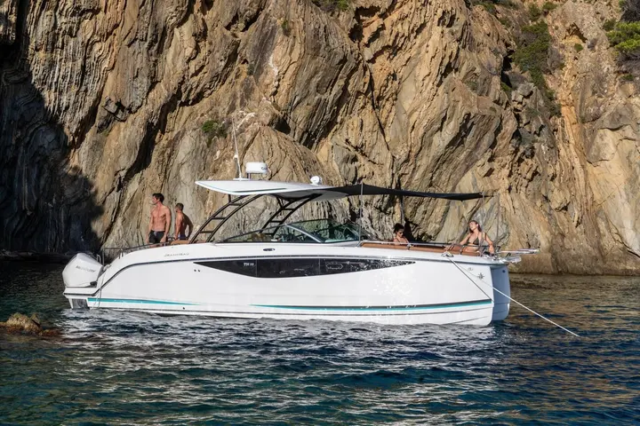 Jeanneau TH33: white hull at anchor beside tall rock cliffs, people on deck under the hardtop and bow sunshade