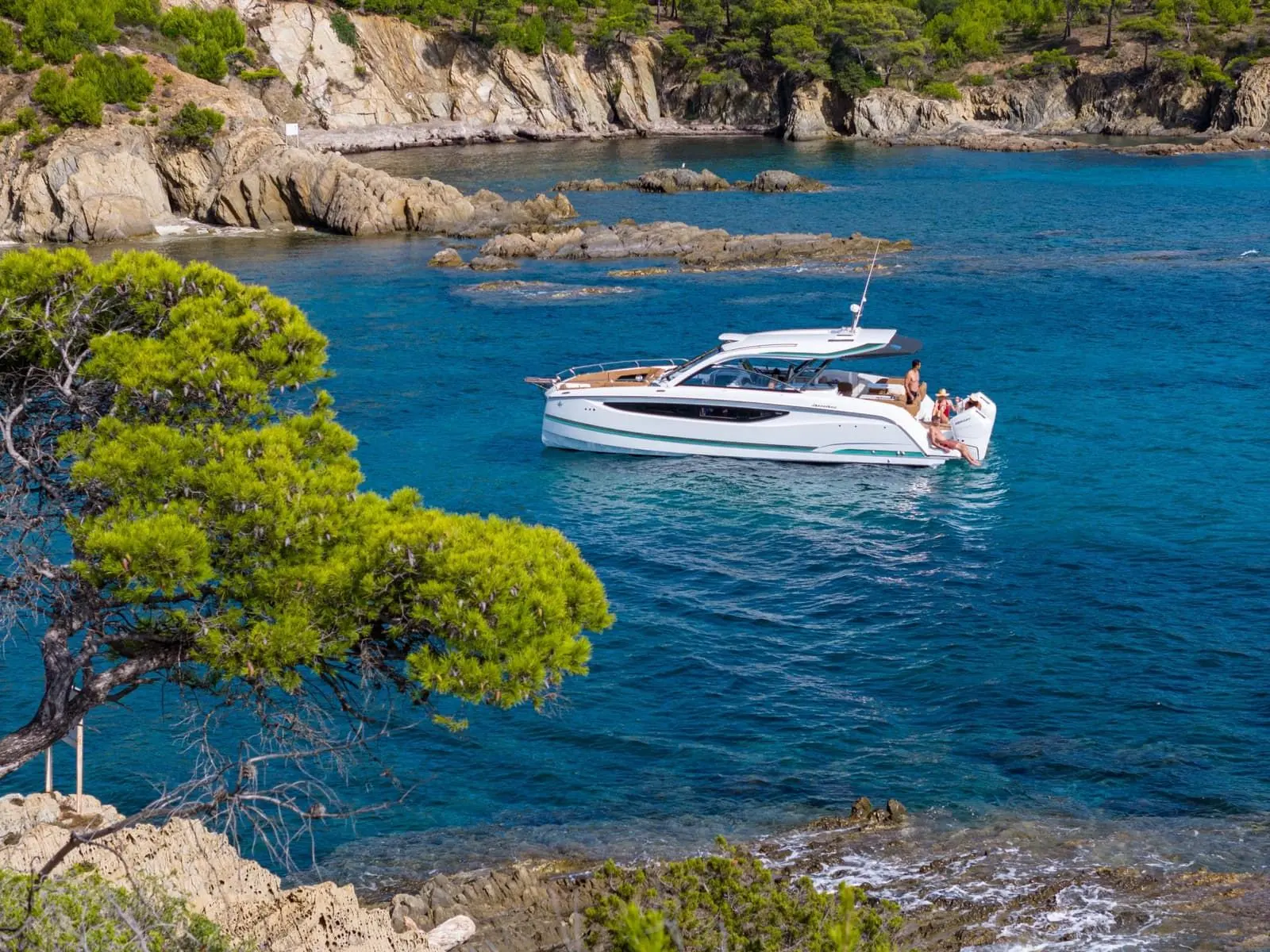 Jeanneau TH38: boat at anchor in a turquoise rocky cove, guests seated aft, pine tree in foreground