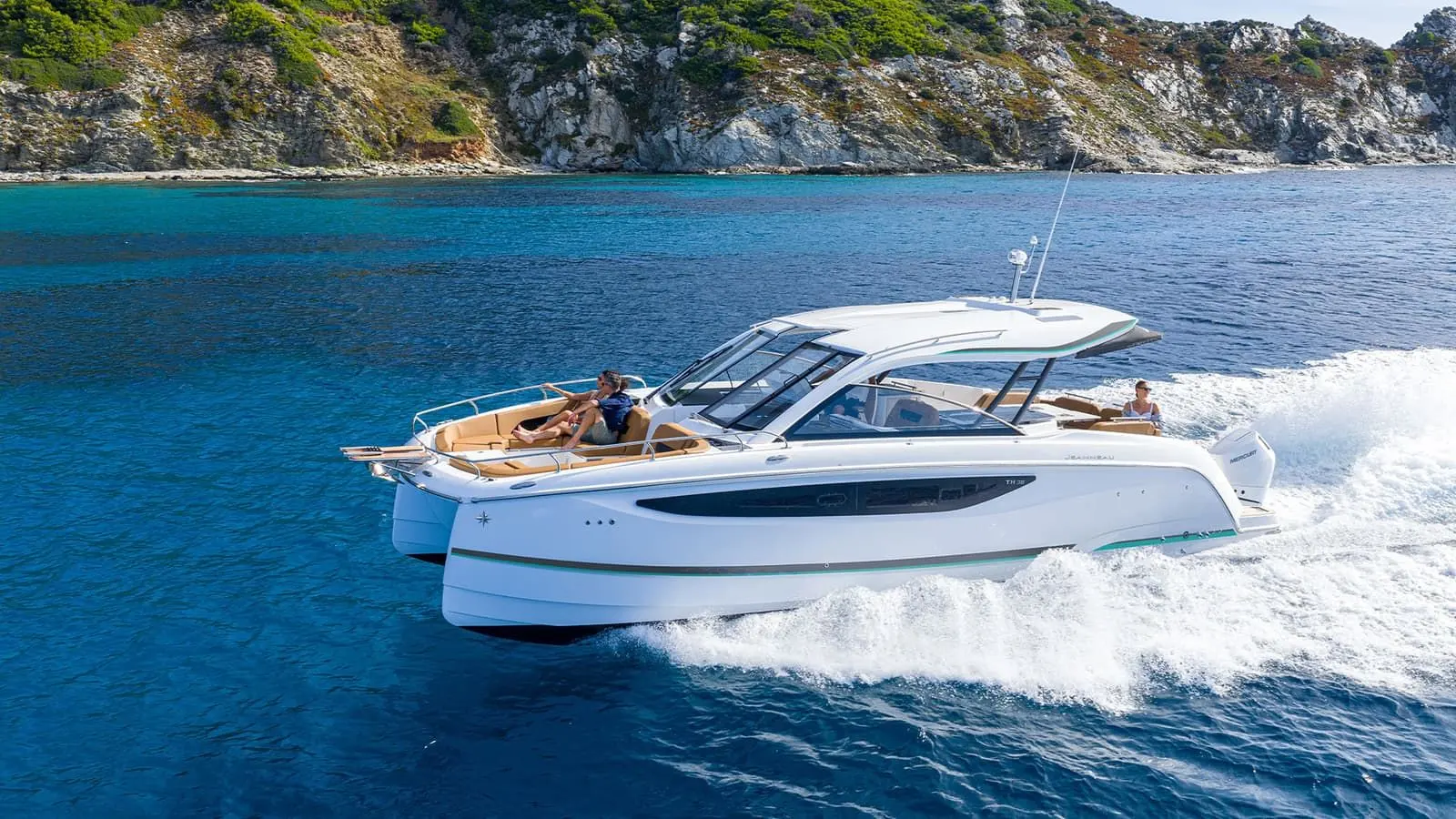 Jeanneau TH38: white motorboat under way at speed, people relaxing on foredeck sunbed beneath rocky coastline