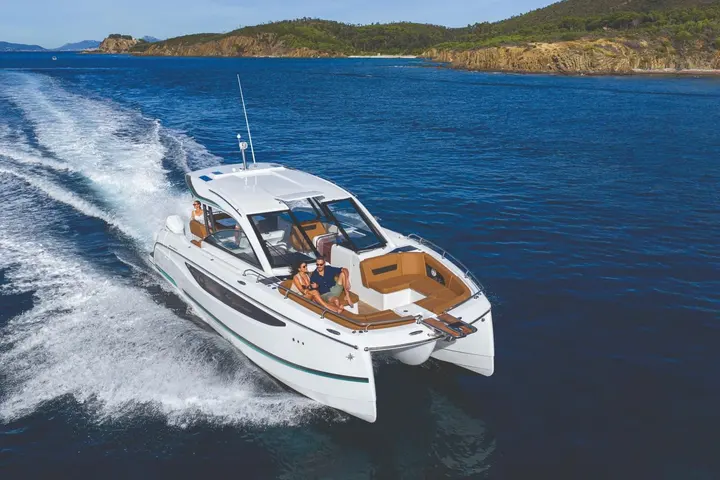 Jeanneau TH38: white power catamaran under way leaving a wake, couple seated in tan bow lounge near rocky coast