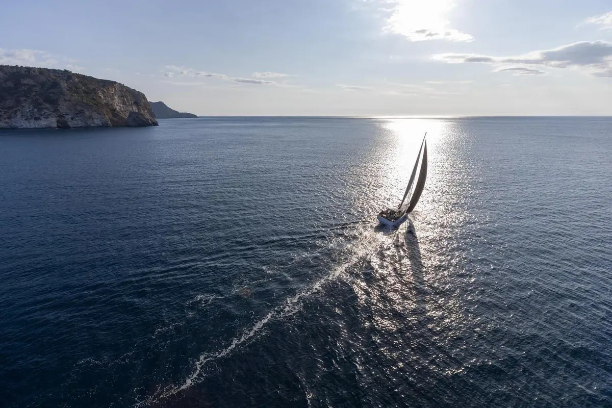 Jeanneau Yachts 55: aerial view of the yacht under sail towards the sun, leaving a long wake near rocky headland