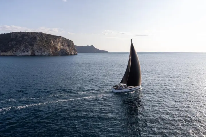 Jeanneau Yachts 55: yacht under sail with dark mainsail and headsail, white hull and wake, cliffs behind