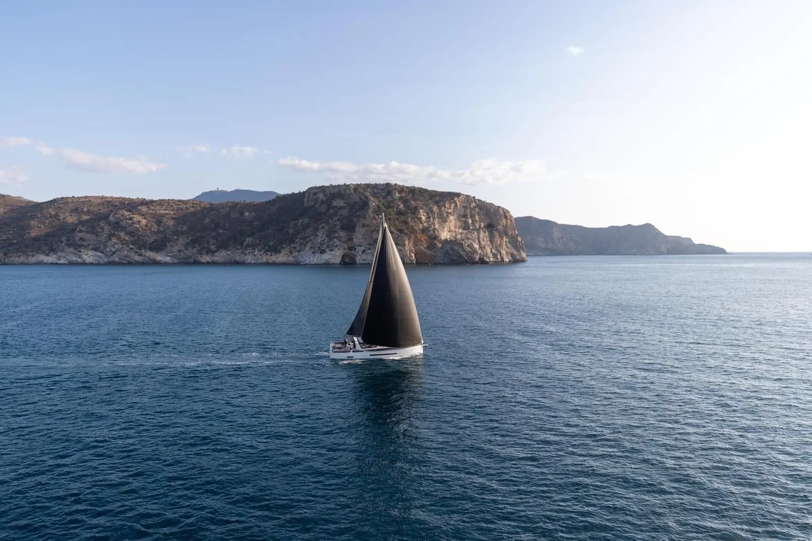 Jeanneau Yachts 55: side view under sail with dark sails on calm blue water in front of a rocky coastline