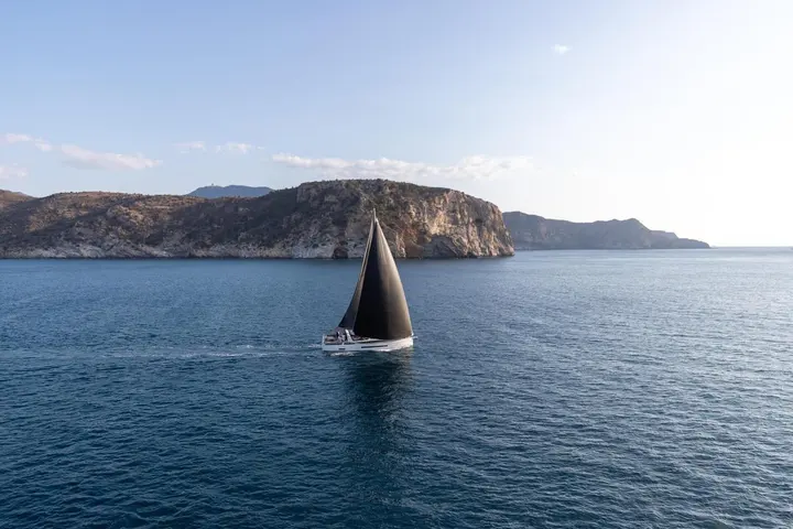 Jeanneau Yachts 55: side view under sail with dark sails on calm blue water in front of a rocky coastline