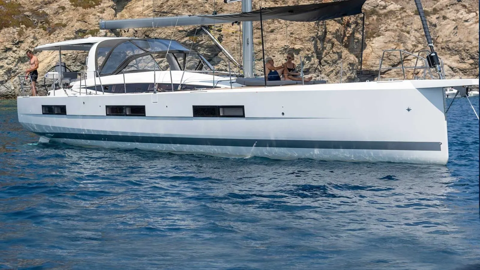 Jeanneau Yachts 60: side view at anchor near rocky shore, white hull, people relaxing on deck and cockpit