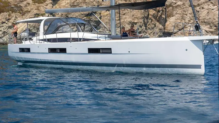 Jeanneau Yachts 60: side view at anchor near rocky shore, white hull, people relaxing on deck and cockpit