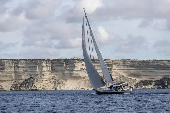 Jeanneau Yachts 65: dark-hulled yacht under sail heeling past tall white limestone cliffs under a cloudy sky