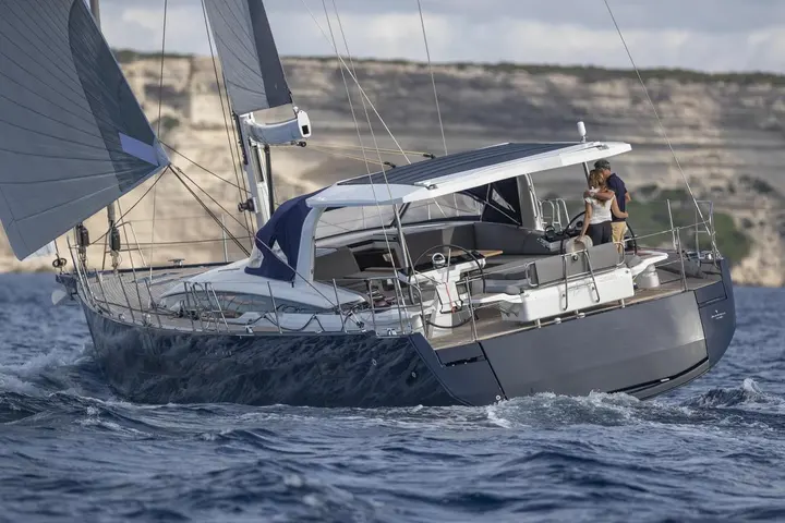 Jeanneau Yachts 65: stern quarter view under sail, couple at the helm beneath a hard cockpit roof, cliffs behind
