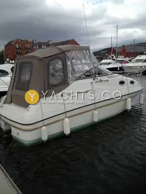Regal 242 Commodore: starboard side afloat with beige camper covers fitted over the cockpit, marina flats behind