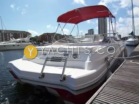 Rinker 210 MTX: stern view showing bathing ladder, full-width sunpad and red bimini, with marina flats behind