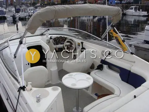 Rinker 232: cockpit under cream bimini with helm, bucket seats, pedestal table and blue-trimmed seating
