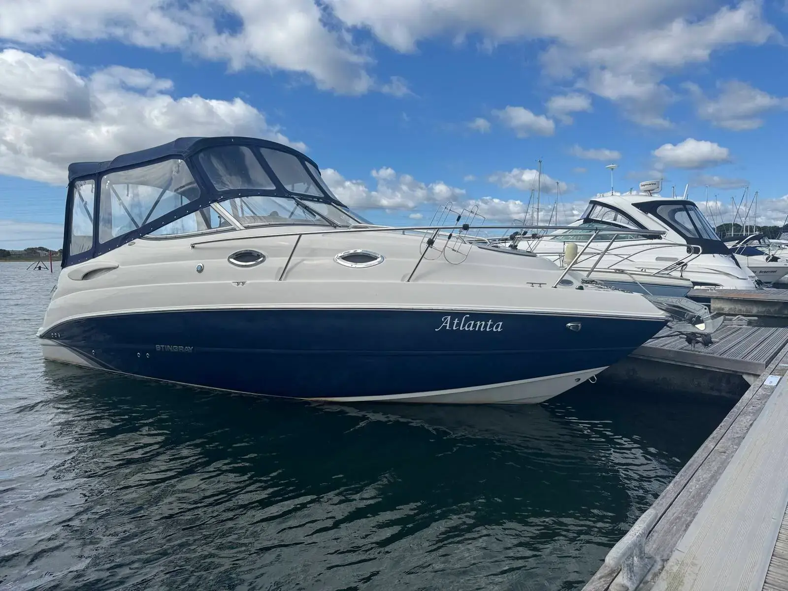Stingray 250 CS: navy and white hull with name on the side, camper cover fitted, moored at a marina pontoon