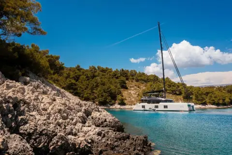 Sunreef Eco 70: catamaran at anchor in a clear bay, seen past rocky shoreline and pine trees
