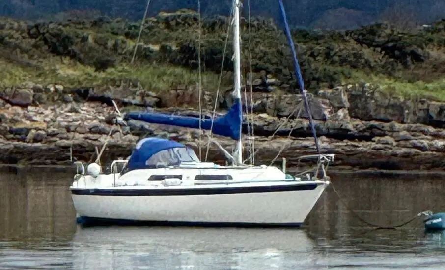 Westerly Merlin 28: white hull at anchor with blue sail covers, rocky wooded shoreline behind